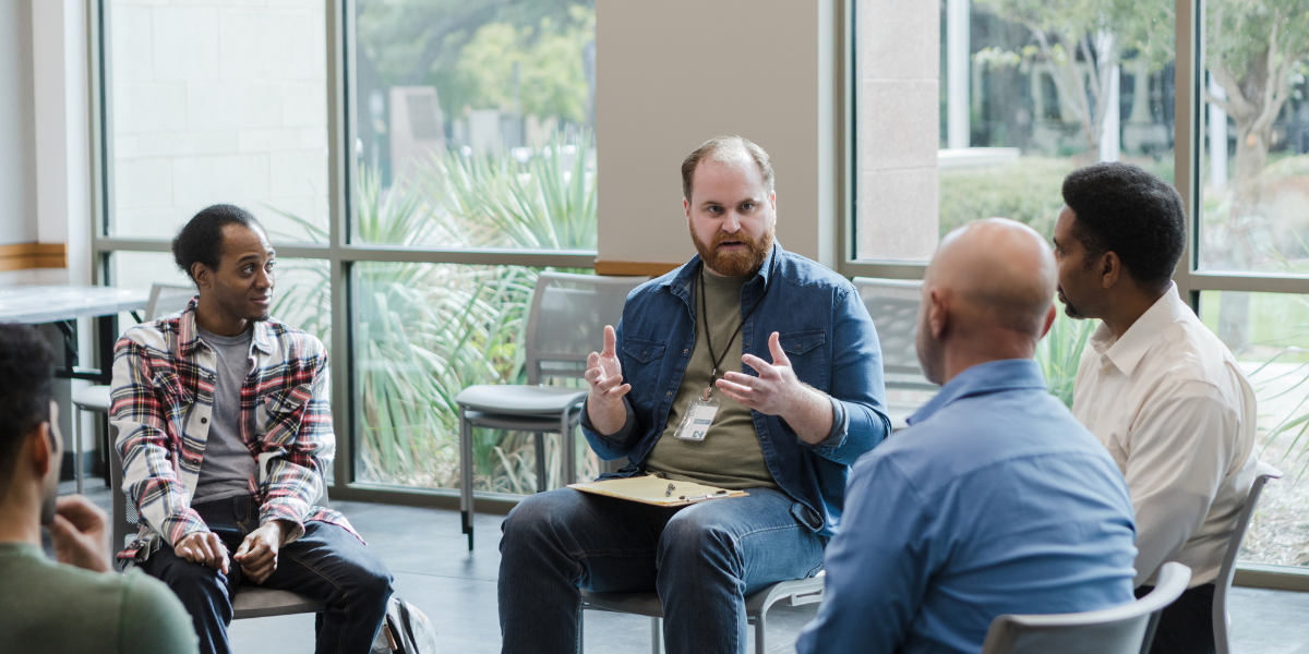 Men sitting on chairs around a circle in a group setting, having a discussion