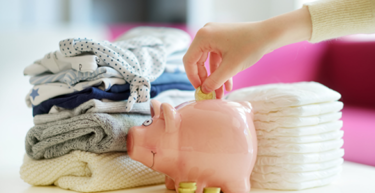 piles of neatly folded nappies and baby clothes with a piggy bank in front and a hand putting money into the piggy bank.