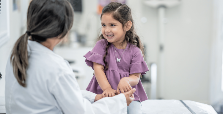 young child, a girl is sitting at the doctor's room on the bed smiling and looking at the female doctor