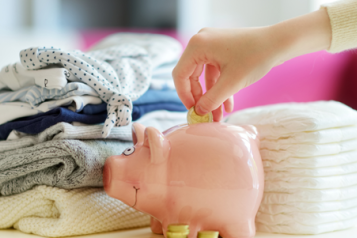 piles of neatly folded nappies and baby clothes with a piggy bank in front and a hand putting money into the piggy bank.