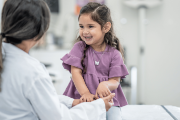 young child, a girl is sitting at the doctor's room on the bed smiling and looking at the female doctor