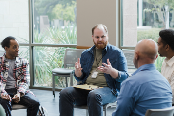 Men sitting on chairs around a circle in a group setting, having a discussion